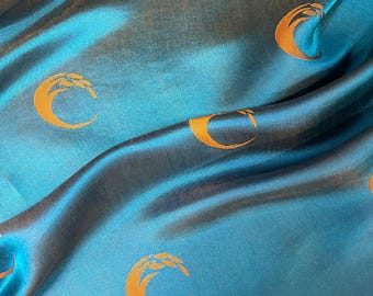 May include: Close-up of a teal fabric with a satin sheen. The fabric is patterned with repeating gold crescent moon shapes. The folds in the fabric create shadows and highlights, adding depth to the design.