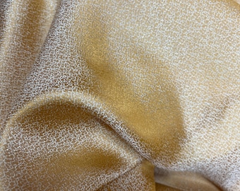 May include: Close-up of a golden fabric with a subtle, textured pattern. The material has a shimmering quality, with highlights and shadows creating depth. The fabric appears to be draped, showcasing its fluidity and drape.