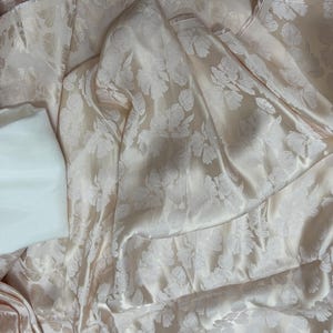 Könnte beinhalten: Ein blassrosa Satin-Stoff mit einem weißen Blumenmuster. Der Stoff ist drapiert und gefaltet, wodurch die Textur und der Glanz des Materials sichtbar werden.