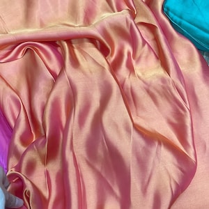 May include: Close-up of draped fabric in shades of pink, peach, and teal. The fabric has a shiny, satin-like texture, with folds and shadows creating depth. The colours blend and shift, giving a gradient effect.