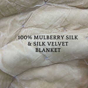 Può includere: Primo piano di una coperta in velluto di seta color crema. La coperta ha un design trapuntato con piccole cuciture azzurre. Il testo "100% MULBERRY SILK & SILK VELVET BLANKET" è stampato.