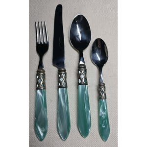Puede incluir: Un juego de cubiertos de cuatro piezas con tenedor, cuchillo y dos cucharas. Los mangos son de color verde marmoleado con una banda decorativa plateada. Los utensilios están colocados sobre una superficie neutra.