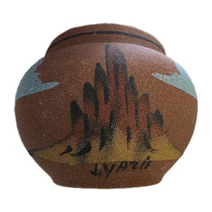 Puede incluir: Una olla de cerámica marrón con una superficie texturizada. La olla presenta un diseño de paisaje pintado con acentos negros, rojos y amarillos. El diseño incluye una cordillera y nubes. La firma del artista, "J. Yazzie", es visible.