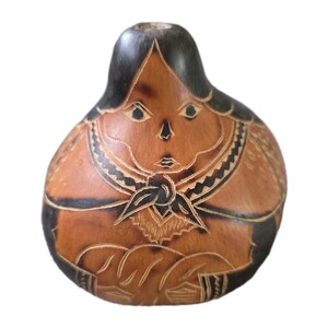 Può includere: Oggetto decorativo a forma di zucca con un volto e una figura scolpiti. Il volto è marrone con capelli e lineamenti neri. La figura ha un colletto e braccia dettagliati, tutti scolpiti sulla superficie della zucca.
