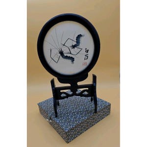 May include: A black framed circular artwork featuring a minimalist ink painting of two shrimp. The artwork is mounted on a black wooden stand, which rests on a patterned box. The artwork includes Chinese characters.