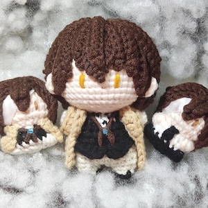 May include: Crocheted doll of a character with brown hair, yellow eyes, and a black vest over a white shirt. The doll has a tan coat and is flanked by two crocheted head pieces. The doll is on a white surface.