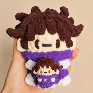 Puede incluir: Muñeca de ganchillo hecha a mano con un diseño de dibujos animados. La muñeca tiene cabello castaño texturizado, una cara color crema y un atuendo morado. Una versión más pequeña de la muñeca está unida al frente. La muñeca se sostiene en una mano.