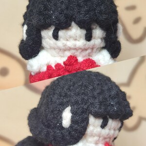 May include: A crocheted doll with black hair, a white face, and a red sweater. The doll is wearing a white scarf.