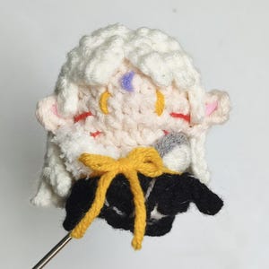May include: A crocheted plush toy with white hair, pink ears, and a yellow bow. The toy is wearing a black and white outfit and is holding a microphone.