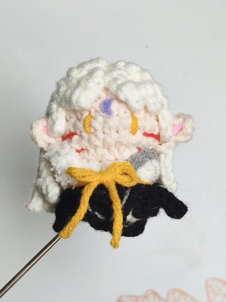 May include: A crocheted plush toy with white hair, pink ears, and a yellow bow. The toy is wearing a black and white outfit and is holding a microphone.