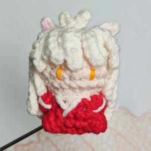 May include: A crocheted plush toy of a white-haired character with red clothing and yellow eyes. The toy is made with a soft, textured yarn and has a cute, cartoonish appearance.