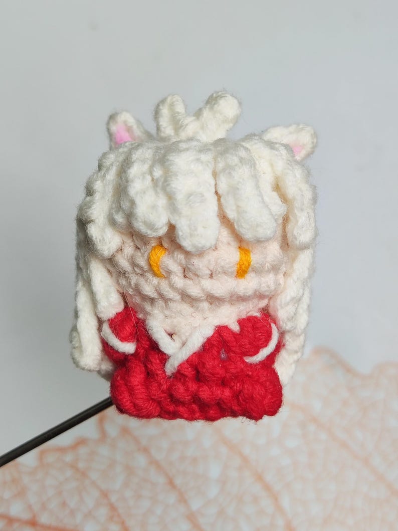May include: A crocheted plush toy of a white-haired character with red clothing and yellow eyes. The toy is made with a soft, textured yarn and has a cute, cartoonish appearance.