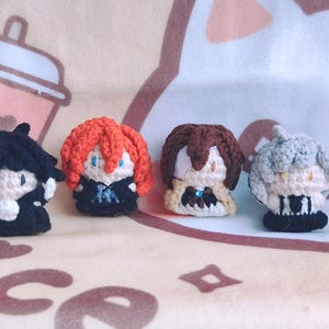 May include: Four crocheted plushie figures, each with a different hair colour and outfit. The figures are standing on a light brown fuzzy surface.