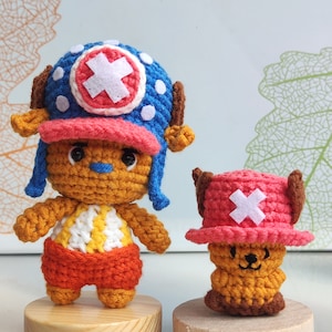 Puede incluir: Dos peluches de crochet del personaje Tony Tony Chopper del anime One Piece. El juguete más grande está de pie sobre una base de madera y lleva un sombrero azul y rojo con una cruz blanca. El juguete más pequeño está sentado sobre una base de madera y lleva un sombrero rojo con una cruz blanca.