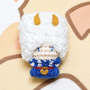 May include: A handmade crochet doll with a white fluffy hat and two yellow horns. The doll has a blue and white outfit with a gold-colored emblem. The doll's face is beige with blue eyes.