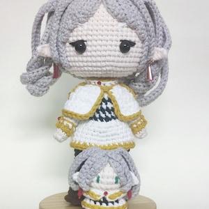 Könnte beinhalten: Zwei gehäkelte Puppen, eine steht auf der anderen. Die größere Puppe hat graues Haar, ein weißes und schwarzes Kleid und rote Ohrringe. Die kleinere Puppe hat graues Haar, ein gelbes und schwarzes Kleid und grüne Augen. Beide Puppen stehen auf einem Holzsockel.