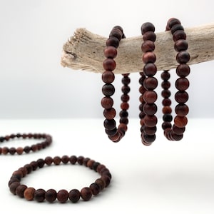 May include: Three brown wooden bead bracelets stacked on a piece of driftwood. The bracelets are made of round beads and are approximately 18 cm in length.