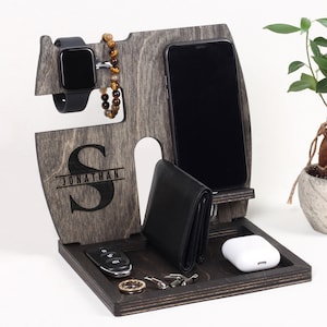 May include: A wooden organizer with a gray finish. The organizer has slots for a phone, watch, and wallet. There is a small tray for keys and other small items. The organizer has the name 'JONATHAN' engraved on the front. The organizer is sitting on a white surface next to a small potted plant.