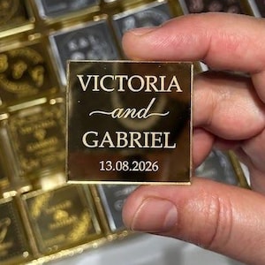 May include: A square gold mirrored acrylic piece featuring the engraved text VICTORIA and GABRIEL with the date 13.08.2026. The piece is held in front of a grid of identical square gold and dark grey mirrored acrylic tiles with various designs.