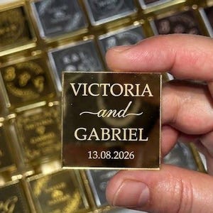 Może przedstawiać: Kwadratowa, złotego koloru płytka akrylowa z wygrawerowanym napisem VICTORIA and GABRIEL oraz datą 13.08.2026. Płytka jest trzymana przed siatką podobnych złotych i ciemnych kwadratowych płytek z różnymi wygrawerowanymi wzorami.