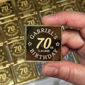 May include: A collection of square gold-colored mirrored acrylic tiles, each engraved with the text GABRIEL'S 70TH BIRTHDAY and the date 11.04.2028. The tiles feature a central number 70, small star accents, and a reflective metallic finish.