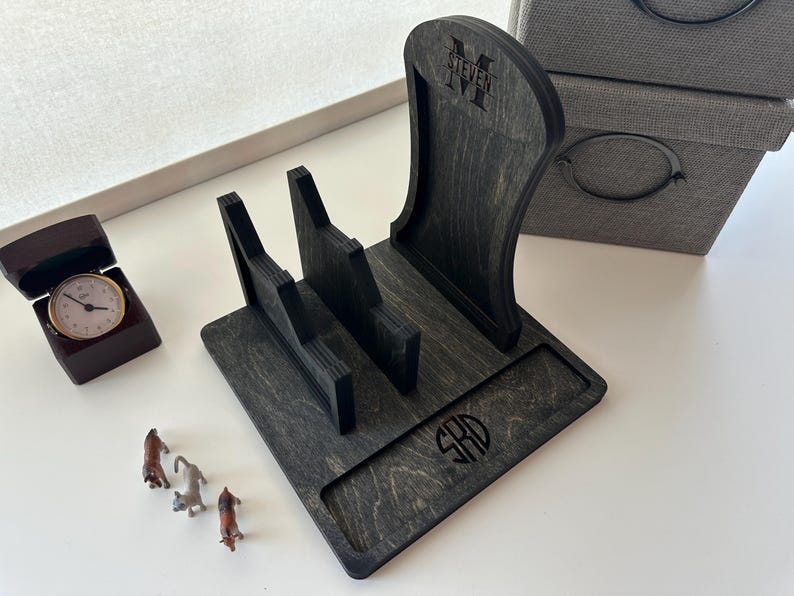May include: A black wooden phone stand with a monogrammed design. The stand has a raised back and a flat front with a small compartment for charging cables. The monogram is "SHD".