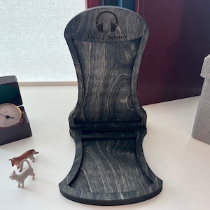 Pode incluir: Um suporte para fones de ouvido de madeira preta com um recorte de fones de ouvido e o nome "David Adams" gravado na frente. O suporte &eacute; feito de madeira com uma mancha cinza escura.