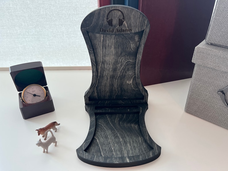 Pode incluir: Um suporte para fones de ouvido de madeira preta com um recorte de fones de ouvido e o nome "David Adams" gravado na frente. O suporte &eacute; feito de madeira com uma mancha cinza escura.