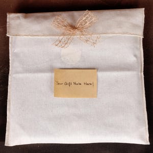 May include: A white fabric gift bag with a beige bow and a small, rectangular card that reads "Your Gift Note Here!". The bag is closed at the top and sits on a dark brown surface.