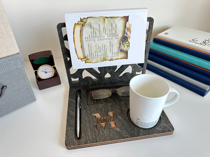 May include: A dark wood desk organiser with a personalised name, "James", engraved on the front. It features a built-in pen holder, a slot for glasses, and a cup holder. The organiser is displayed on a white surface with a stack of books and a small clock.