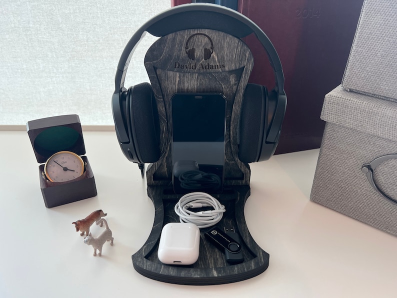 Pode incluir: Um suporte para fones de ouvido de madeira com acabamento em madeira preto e cinza. O suporte tem um suporte para telefone, um espa&ccedil;o para fones de ouvido e uma unidade USB. O suporte &eacute; personalizado com o nome "David Adams".