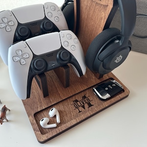 May include: Wooden gaming accessory organizer with two white and black game controllers, a black headset, a car key, and white earbuds. The organizer has a tray with the name "OLIVER" engraved.
