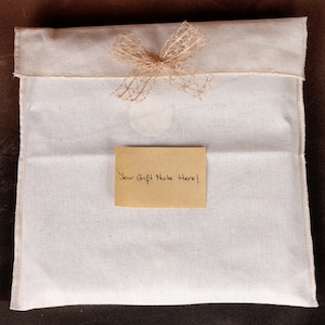 May include: A gift wrapped in a white fabric bag, secured with a natural raffia bow. A small brown card with the handwritten text "Your Gift Note Here!" is attached. The words "GIFT WRAPPING AVAILABLE" are printed vertically on the left.