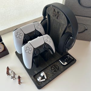 May include: A wooden gaming console stand with a black headset, two white controllers, and a small black remote. The stand is personalized with the initials "SHD".