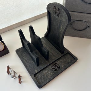 May include: A dark gray wooden phone stand with a curved back and two slots. The stand has the name "STEVEN" and initials engraved on it. A small watch in a brown box and miniature dog figurines are also present.
