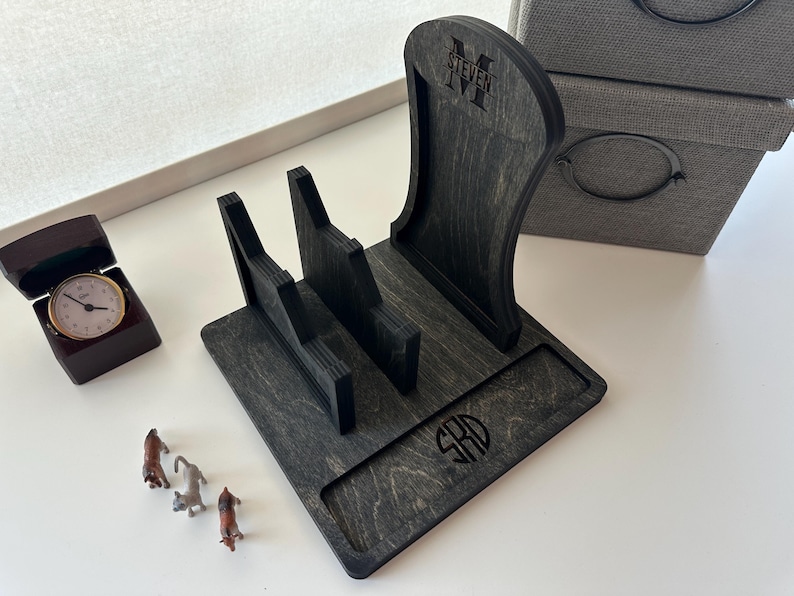 May include: A dark gray wooden phone stand with a curved back and two slots. The stand has the name "STEVEN" and initials engraved on it. A small watch in a brown box and miniature dog figurines are also present.