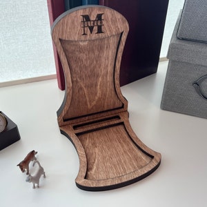 Pode incluir: Um suporte de telefone de madeira com acabamento marrom escuro. O suporte tem um recorte para um telefone e uma se&ccedil;&atilde;o elevada para o telefone descansar. O suporte &eacute; personalizado com o nome "Oliver" e a inicial "M" em letras pretas.