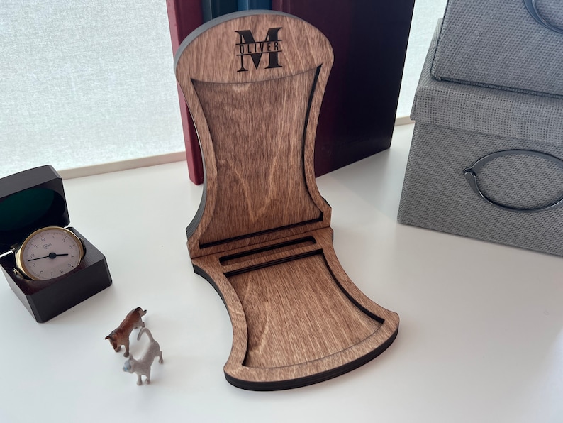 Pode incluir: Um suporte de telefone de madeira com acabamento marrom escuro. O suporte tem um recorte para um telefone e uma se&ccedil;&atilde;o elevada para o telefone descansar. O suporte &eacute; personalizado com o nome "Oliver" e a inicial "M" em letras pretas.