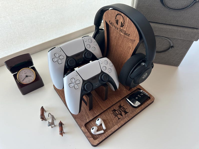May include: A wooden stand with a personalized name, "Oliver Miller",  holds two white wireless game controllers, a pair of black headphones, and a small black remote. The stand is made of dark wood and has a slot for each item.