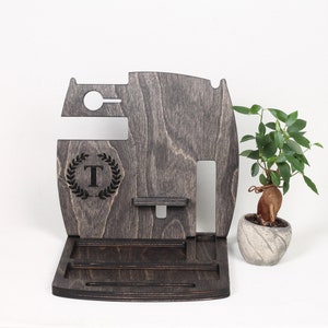 May include: A dark grey wooden desk organiser with compartments for a phone, watch, and accessories. Features a monogrammed "T" within a laurel wreath. A small potted plant is to the right.