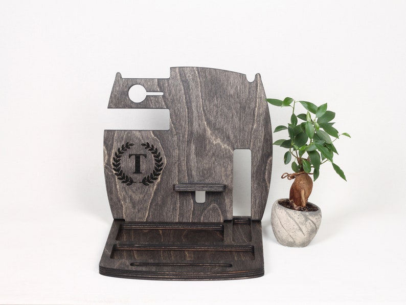 May include: A dark grey wooden desk organiser with compartments for a phone, watch, and accessories. Features a monogrammed "T" within a laurel wreath. A small potted plant is to the right.
