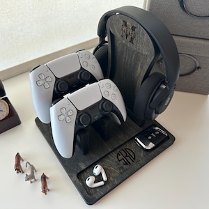 May include: A dark gray wooden gaming station holds two white game controllers, a black headset, and a car key fob. The station is personalized with initials and a name. A small clock and figurines are also present.