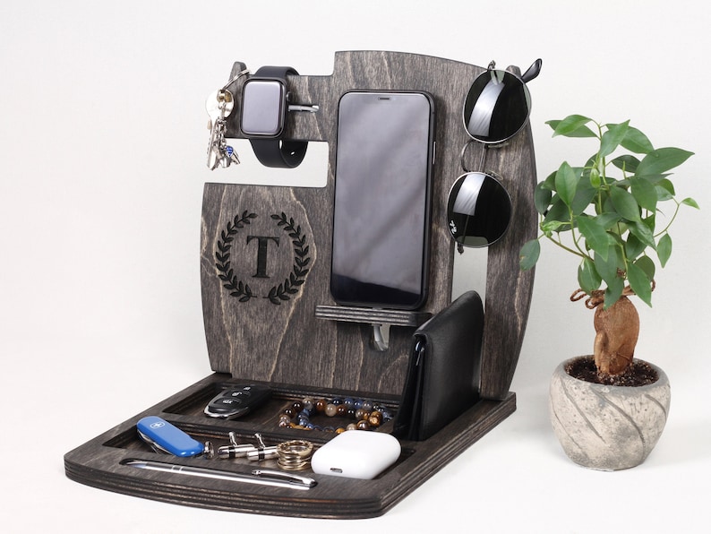May include: A wooden organizer with a gray finish, featuring slots for a phone, watch, and sunglasses. It also has a compartment for keys, a pen, and other small items. The organizer has a personalized inscription with the letter "T" in a laurel wreath.