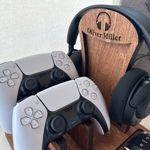 May include: A wooden stand with a personalized nameplate that reads "Oliver Miller" holds a pair of black headphones and two white wireless game controllers.
