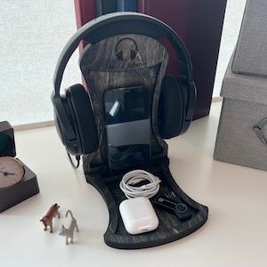 Pode incluir: Um suporte para auscultadores de madeira com acabamento cinzento. O suporte tem um suporte para telem&oacute;vel e um espa&ccedil;o para cabos de carregamento. O suporte &eacute; personalizado com o nome "David Adams".