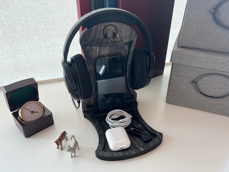 Pode incluir: Um suporte para auscultadores de madeira com acabamento cinzento. O suporte tem um suporte para telem&oacute;vel e um espa&ccedil;o para cabos de carregamento. O suporte &eacute; personalizado com o nome "David Adams".