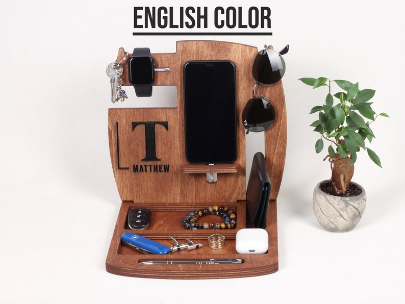 May include: A wooden organiser with spaces for a phone, watch, keys, sunglasses, wallet, and other accessories. The organiser is engraved with the name "Matthew" and the initial "T". A small plant in a decorative pot is on the right.