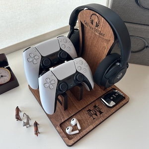 May include: A wooden stand with a personalized name, "Oliver Miller",  holds two white wireless game controllers, a pair of black headphones, and a small black remote. The stand is made of dark wood and has a slot for each item.