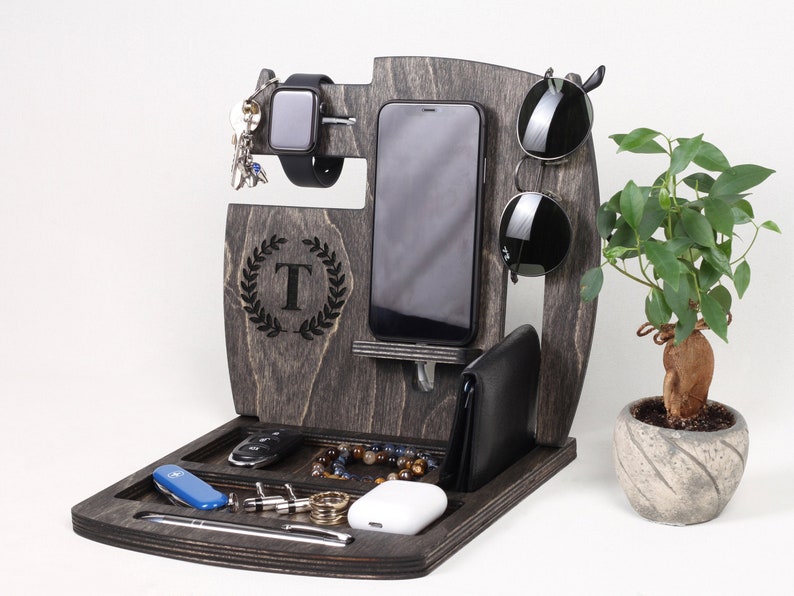 May include: A wooden desk organizer in a dark gray finish, featuring compartments for a phone, wallet, watch, keys, and accessories. The organizer has a laurel wreath with the letter "T" engraved on it. Sunglasses and a small plant are also visible.