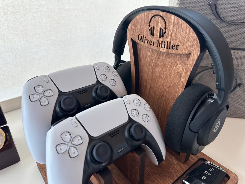 May include: Wooden headphone stand with the name "Oliver Miller" engraved, holding a pair of black headphones. Two white and black game controllers are in front of the stand. A car key fob is visible.
